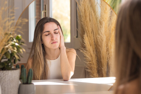 Young woman with eyes closed sitting in front of mirror with headache
