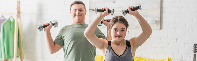 Teenager with down syndrome training with dumbbells near friend at home, banner.