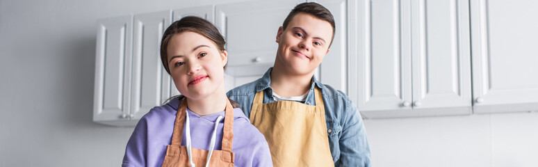 Positive couple with down syndrome in aprons looking at camera in kitchen, banner.