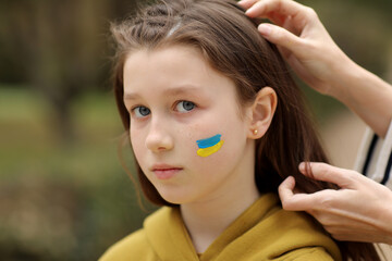 Close up of caring young mother braids hair sad little daughter with flag on face outdoors. Peace, no war, help, stop russian aggression. Invasion of Russia in Ukraine