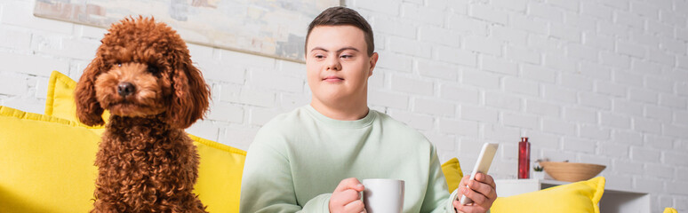 Teen boy with down syndrome holding cup and smartphone near poodle at home, banner.