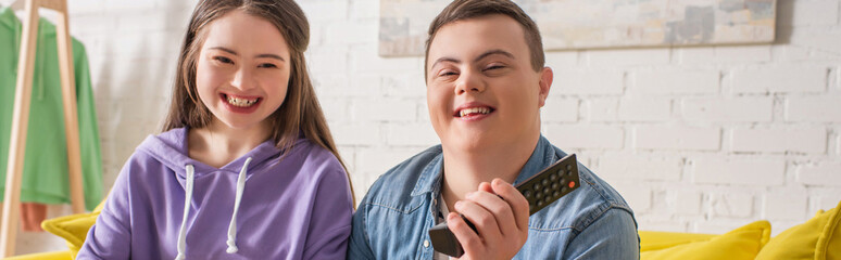 Positive teenagers with down syndrome holding remote controller at home, banner.