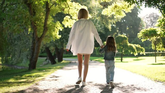 Back view happy young mother and little daughter holding hands walking in sunrays in slow motion outdoors. Wide shot relaxed Caucasian woman and girl enjoying leisure in park strolling