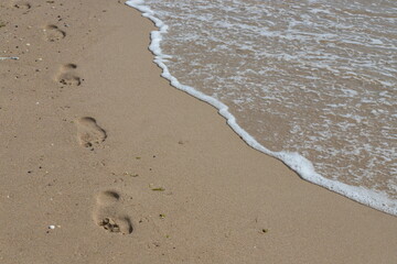 Footprints in the sand beach. Footprints in the sand against a sea wave. Footprints on a sunny day with golden sand, beach, wave and footsteps