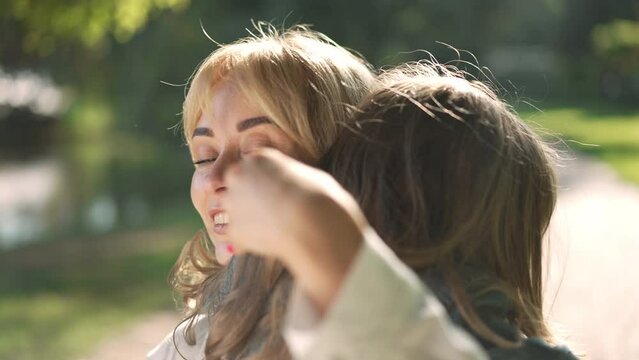 Portrait of beautiful Caucasian young woman hugging cheerful girl looking at camera smiling. Happy mother embracing little daughter posing in slow motion in sunshine in spring summer park