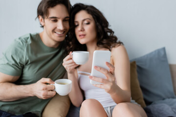 blurred couple with cups looking at smartphone in bedroom.