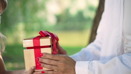 Close-up female hands passing gift box to little girl in sunny park. Unrecognizable Caucasian young woman giving present for daughter on holiday picnic outdoors in slow motion. Celebration concept