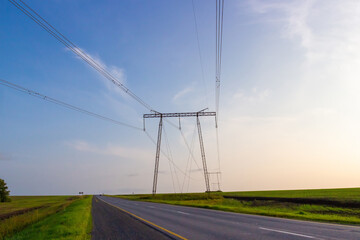High electricity power lines crossing the rural road and green fields during summer sunset. Air...