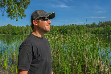 Tanned man in black t-shirt, cap and sunglasses looking deep at beautiful green nature near forest lake