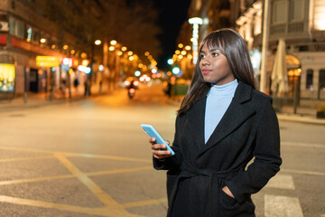 Fototapeta premium Young woman standing on street holding her phone in city night
