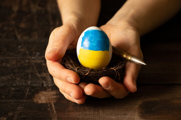 One white chicken egg with a painted flag of Ukraine in a straw basket and a bullet in the hands of...