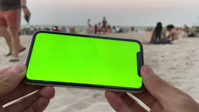 Using phone with green screen chroma key horizontal landscape close up man hand holding smartphone. Blurred tourists relaxing, resting and walking on beach in background - travel and vacation concept