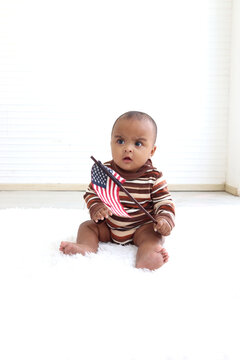 African American Infant Baby Sitting On Floor In Indoor Living Room, Little Kid Holding And Waving American Flag On 4th July To Celebrate National Independence Day, Flag Day Concept.