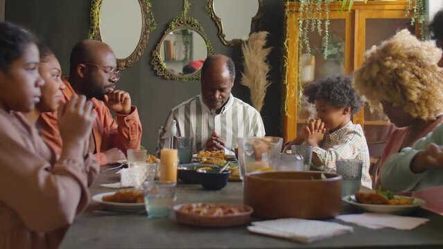 Large African American family listening to father and then praying together with eyes closed before having holiday dinner at home