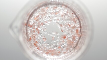 Top view shot of beaker with crystals of brown salt in water on light grey background | Mineral skin care cosmetics formulation concept
