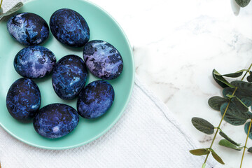 Cosmic-colored eggs lie in a green plate, next to a white waffle towel on a marble table with green leaves. Easter