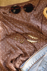 Brown polka dot blouse, vintage mom jeans, straw bag, round sunglasses, gold rings and pearl hair clips. Fashionable outfit on wooden background. Flat lay.