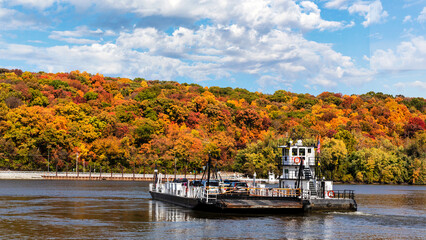 A ferry carries cars across the Mississippi River from Missouri to Illinois and autumn rural countryside landscape with trees with colorful fall leaves on a hillside.