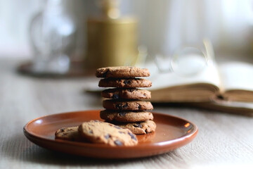 Stack of chocolate chip cookies, open book and reading glasses, lit candle and vase with flowers on the table. Hygge at home. Selective focus.