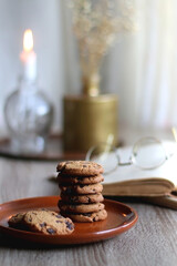 Stack of chocolate chip cookies, open book and reading glasses, lit candle and vase with flowers on the table. Hygge at home. Selective focus.