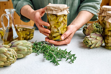 Artichoke hearts marinated with olive oil and herbs. Woman holding a glass jar with pickled...