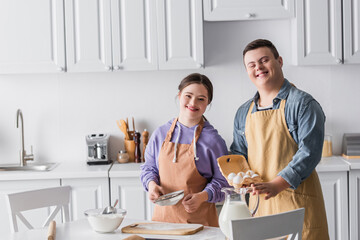 Positive teenagers with down syndrome in aprons cooking and looking at camera in kitchen.