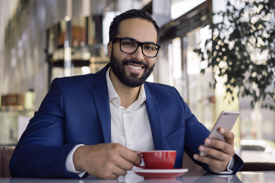 Handsome smiling Arabian businessman using mobile phone drinking coffee in cafe. Portrait of  middle eastern man wearing eyeglasses holding smartphone working online in office. Successful business 
