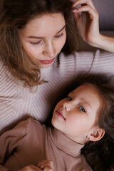 Lovely family mother and daughter embracing and kissing each other indoors. Happy sweet woman with little 6-year-old girl in casual clothers: turtlenecks and jeans sitting on sofa, hugging and smiling