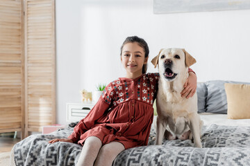 happy brunette girl embracing labrador and smiling at camera while sitting on bed.