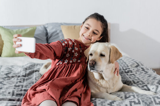 cheerful girl taking selfie on mobile phone with labrador in bedroom.