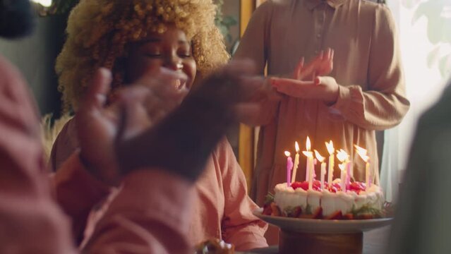 Joyous African American woman covering eyes with hands, then blowing candles on cake and hugging daughter as family clapping hands at home birthday dinner