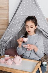 girl drinking tea from toy cup while playing in wigwam.