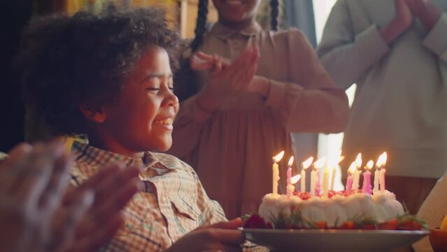 Joyous African American boy blowing candles on birthday cake and happily smiling as family clapping hands for him during home celebration