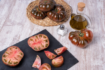 Raf tomato of the black variety, one chopped and the other open in two parts, on a stone plate, on a rustic white table, accompanied by olive oil.