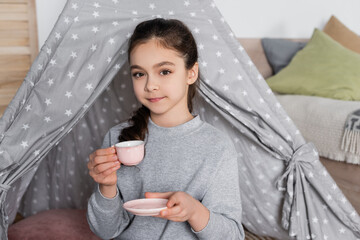 brunette girl with toy cup looking at camera near wigwam.