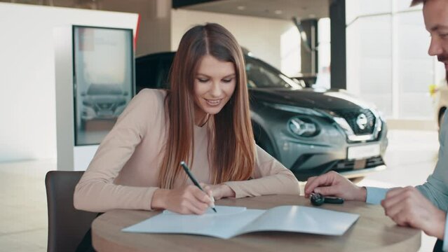 Lady Buying New Car Signing Papers With Dealer Man In Auto Dealership Store. Female customer signing papers at the dealership showroom. Giving the customer the car keys after signing the lease.