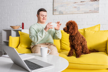 Teenager with down syndrome taking photo of poodle near laptop and cup at home.