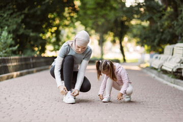 Family walk and fit sport training. Pleasant young arab mother tying shoe laces, while her daughter adjusting her sneakers, sitting on the jogging track in the park at summer morning.