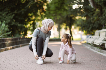 Positive good-looking young mother in hijab, tying laces together with her little daughter, ready to do physical activity outdoors. Family, sport and recreation concept