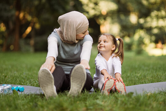 Sport, gymnastics, fitness and physical activity concept, where active smiling Muslim mother having fun with her little daughter, doing stretching exercises on yoga mat outdoors in the park