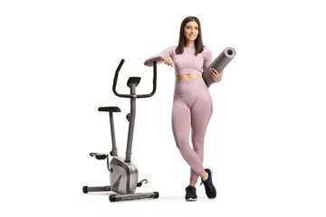 Naklejka na ściany i meble Young woman in sportswear holding an exercise mat next to a stationary bike