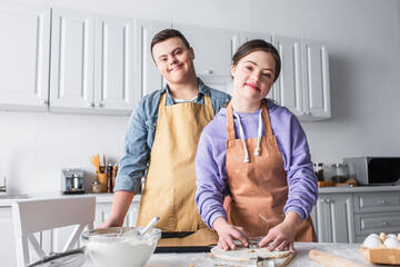 Cheerful couple with down syndrome looking at camera while cooking in kitchen.