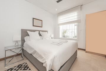 Fototapeta na wymiar Double bed with white pillows and cushions, folded white towels, chrome metal and glass nightstands and a window overlooking a courtyard with plants