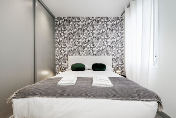bedroom with double bed with white folded towels, white fabric upholstered headboard, green velvet cushions, wall with decorative wallpaper and wardrobe with gray wooden sliding doors