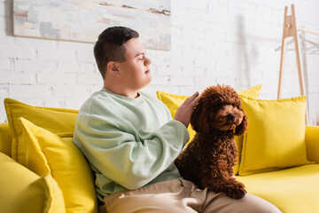 Side view of teen boy with down syndrome petting poodle on couch.