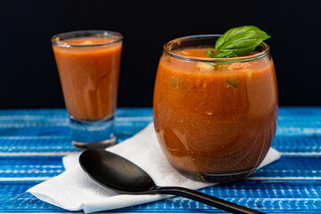 Glass with the refreshing Andalusian Gazpacho on a blue wooden table. One Shot with with Gazpacho too on background. Cold vegetable and organic soup or drink that is drunk in summer or hot days.