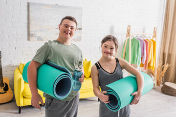 Cheerful teenagers with down syndrome holding fitness mats and sports bottle at home.