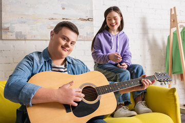 Boy with down syndrome holding acoustic guitar and looking at camera near friend at home.