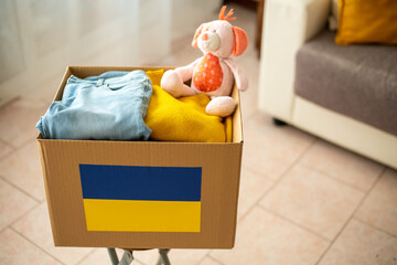 Close up box with Ukrainian flag for humanitarian aid donation with children's things and toys at home on sofa.Stay with Ukraine. Humanitarian aid to Ukrainian refugees. 