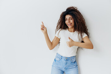 HUGE SALE OFFER CONCEPT. Overjoyed pretty curly Caucasian female in white t-shirt, smile, look at camera, point finger thumb up, gesture Like show copy space. Studio shoot isolated white background